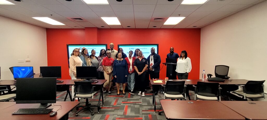 A group of 15 training participants standing in front of a red wall with multiple computer monitors on tables in the foreground.