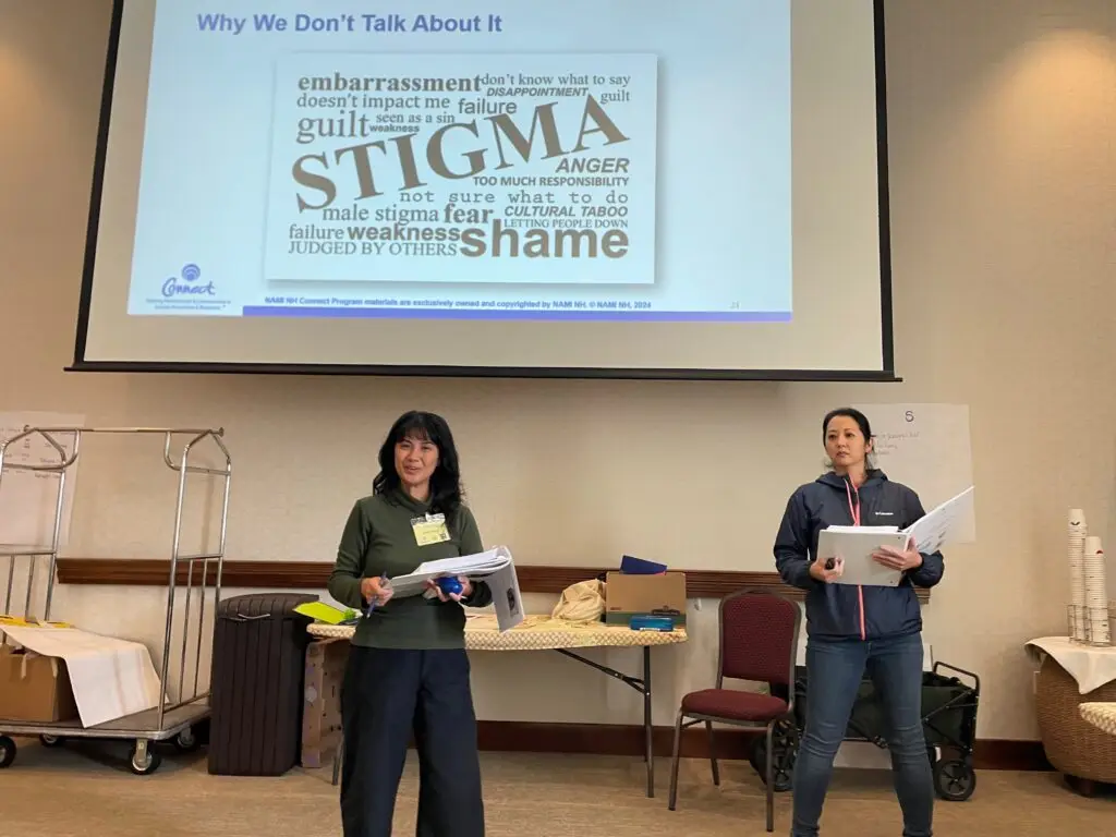 Two women with notebooks presenting in front of a screen displaying "Why we don't talk about it" and works such as stigma, embarrassment, and shame