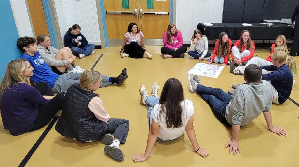 A group of 15 students and teachers sitting in a circle on a gym floor.